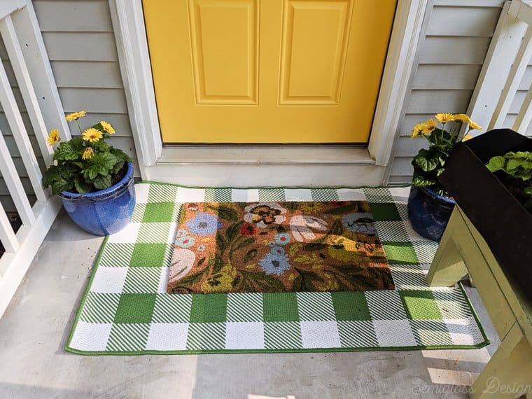 Layered rugs using a buffalo check rug with a floral doormat in front of a yellow front door.